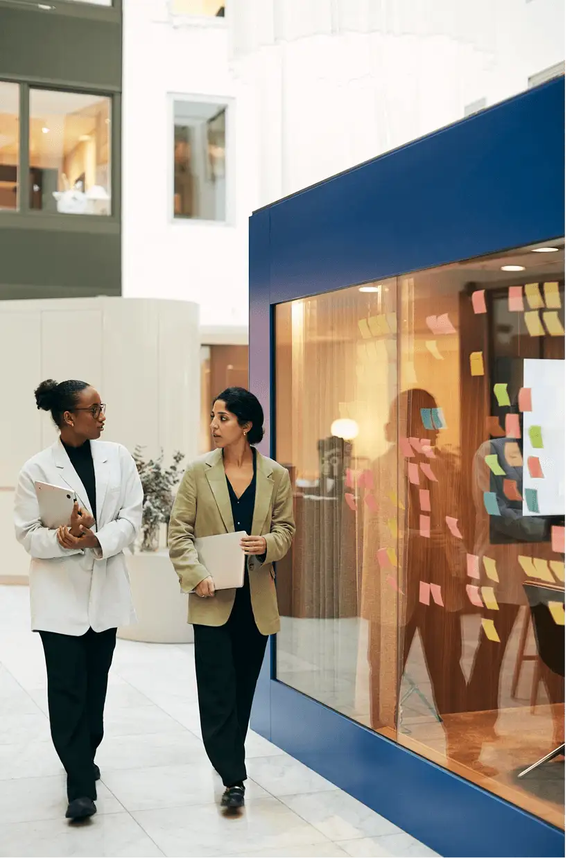 Two women walking in front of a glass wall with sticky notes on it