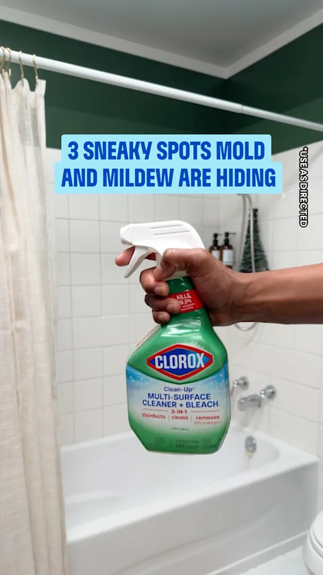 Hand holding Clorox Clean-Up Multi-Surface Cleaner + Bleach spray bottle in a bathroom with white tiles and bathtub.