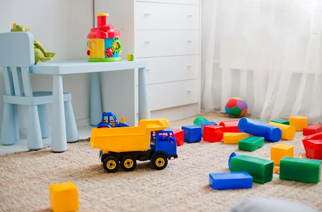 Cuarto de juegos con camión de juguete, bloques coloridos en el suelo, escritorio y silla azules, y cómoda blanca.