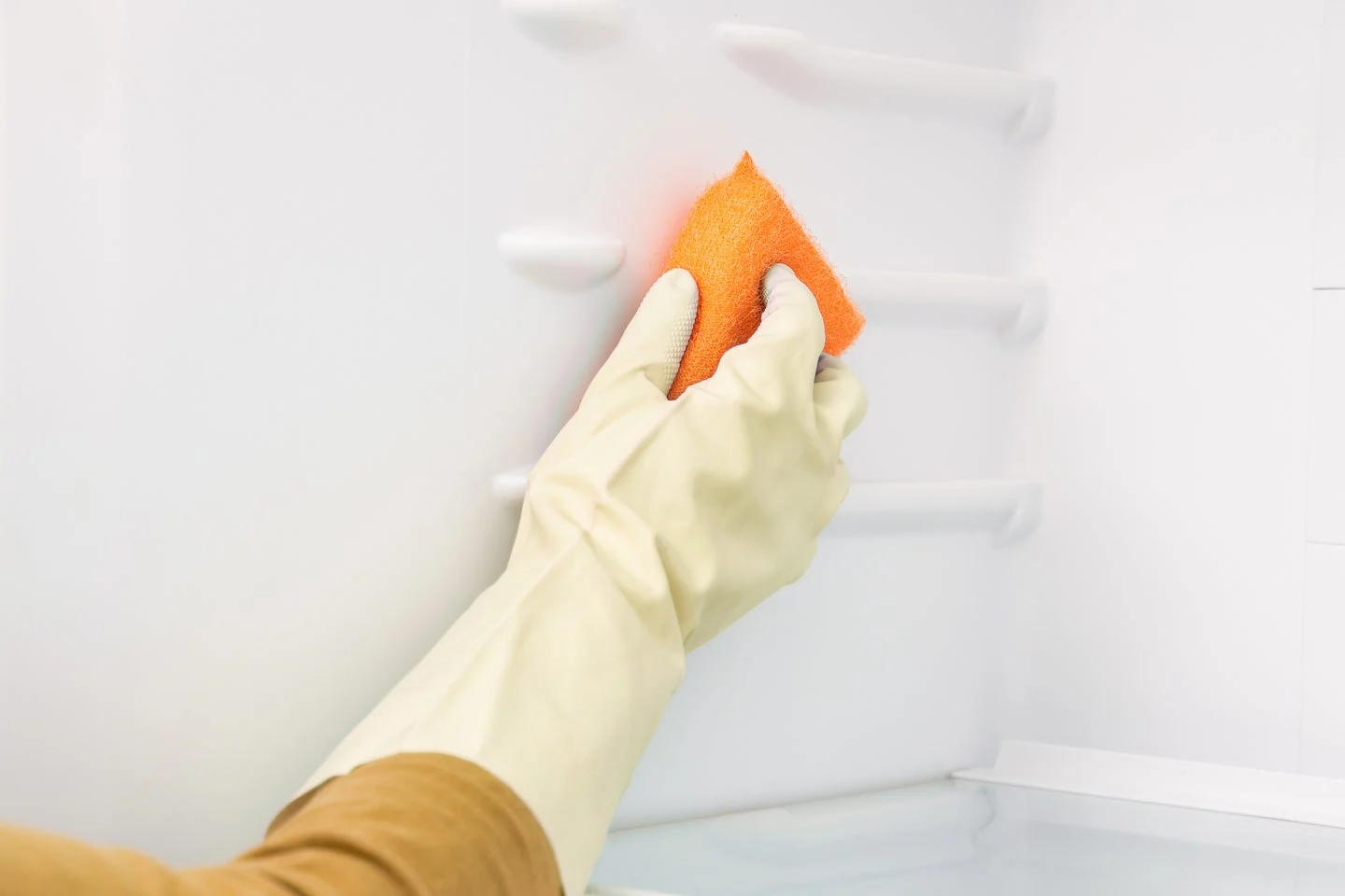 Persona que lleva un guante de limpieza de goma usa una esponja naranja para limpiar el interior blanco de un refrigerador.
