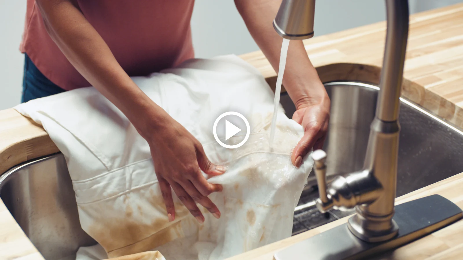 Person washing a stained white piece of clothing in a kitchen sink under running water.