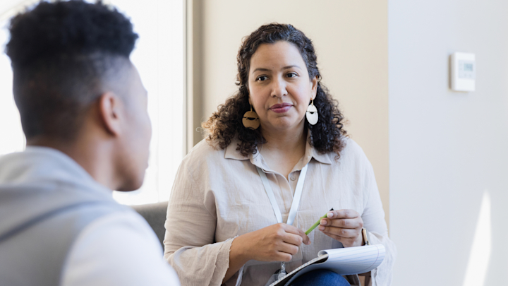 The image shows a therapist talking to a teen patient with a notepad on their lap.